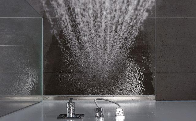 A floor-level shower with glass wall and line drainage is viewed from above. The water flows down from the shower head onto the tiles. 